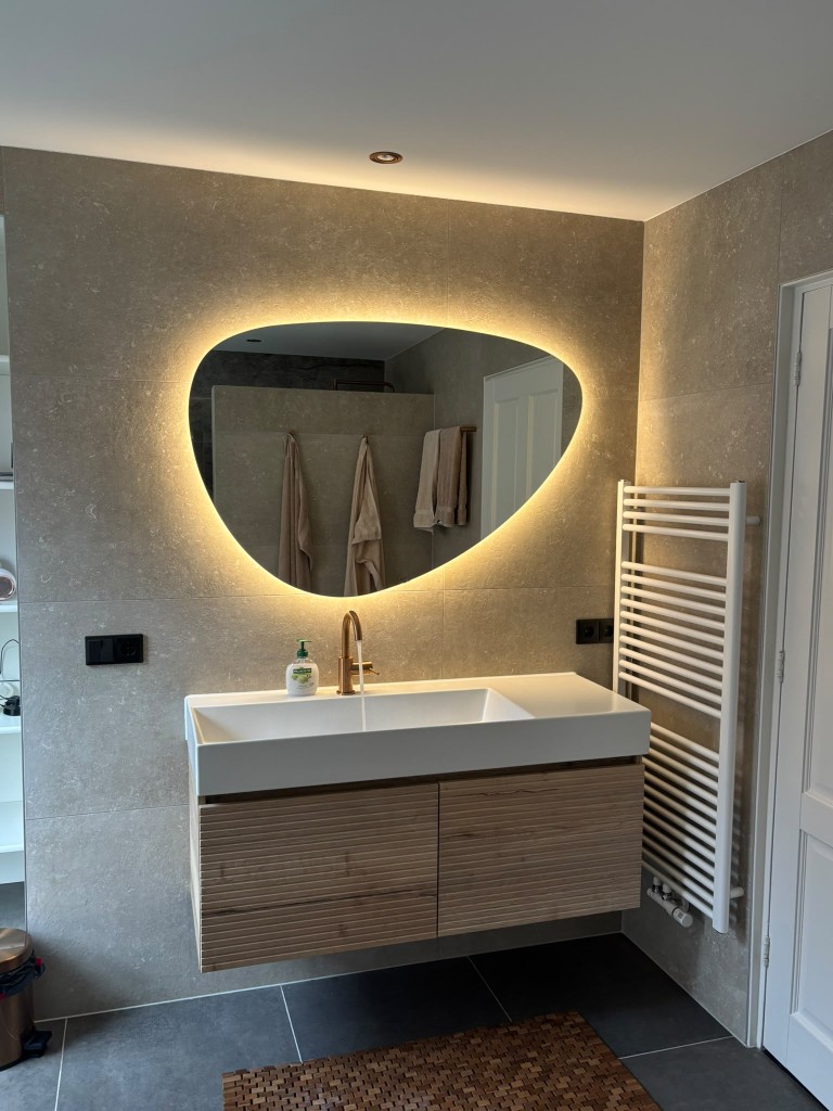 Afgeronde badkamer met organische spiegel, warm licht en een witte wastafel op een houten meubel.